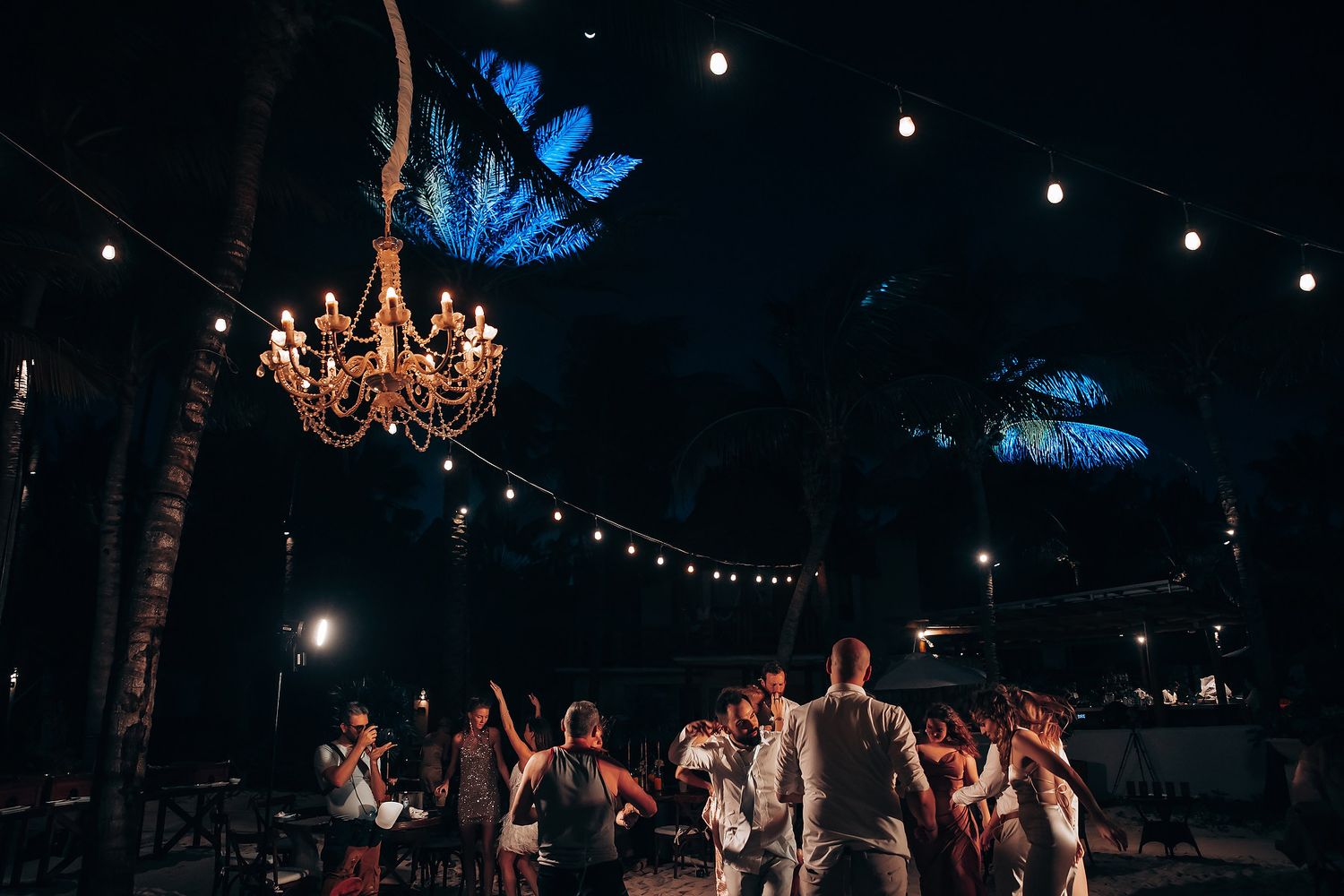 Wedding guests dance under string lights and a crystal chandelier at a beach reception.