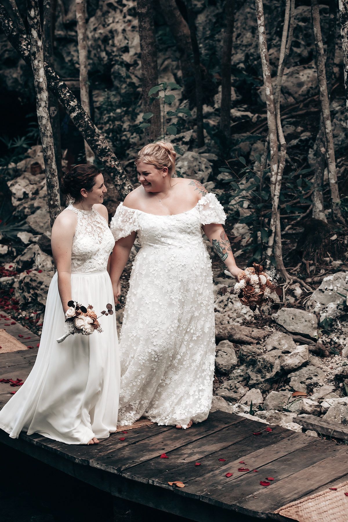 Two women in white wedding dresses stand on a wooden bridge in a lush forest setting.