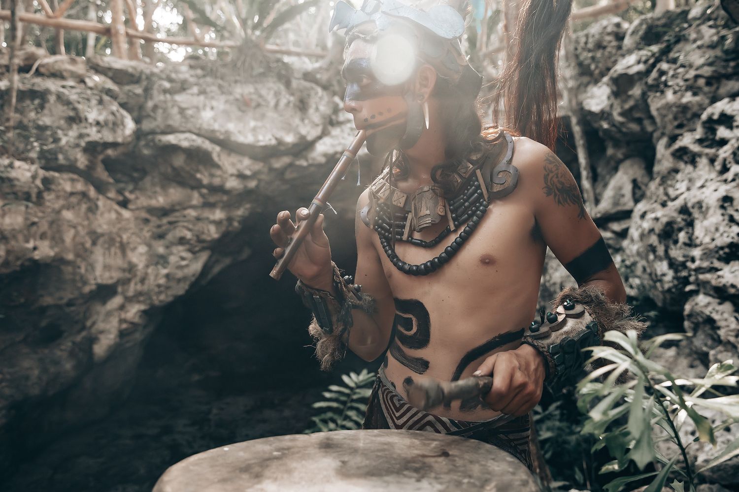 A shirtless tribal man stands in a rocky, forested environment.