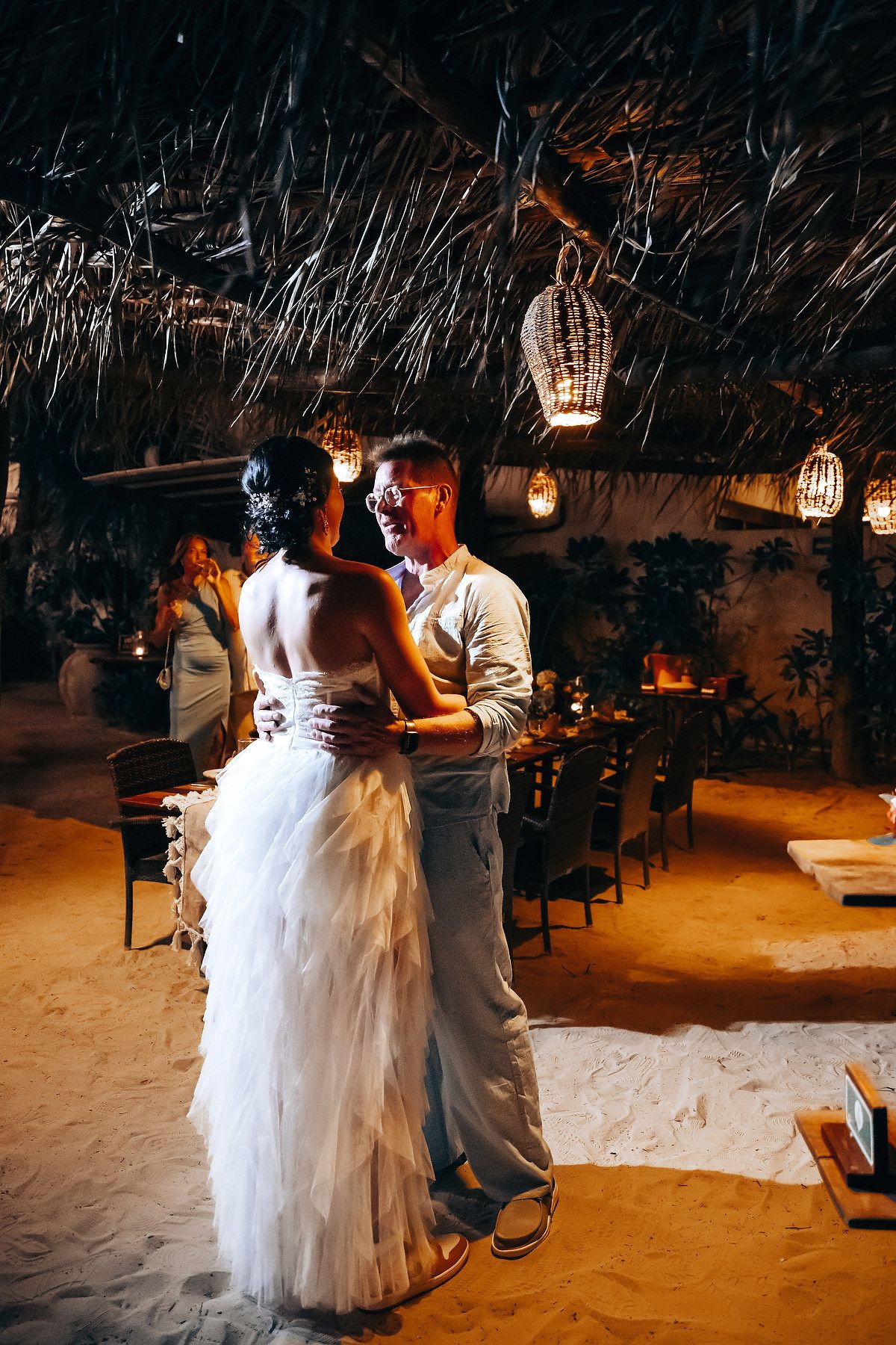 A couple shares a romantic moment at their tropical wedding reception, with hanging lanterns and a fire pit in the background.