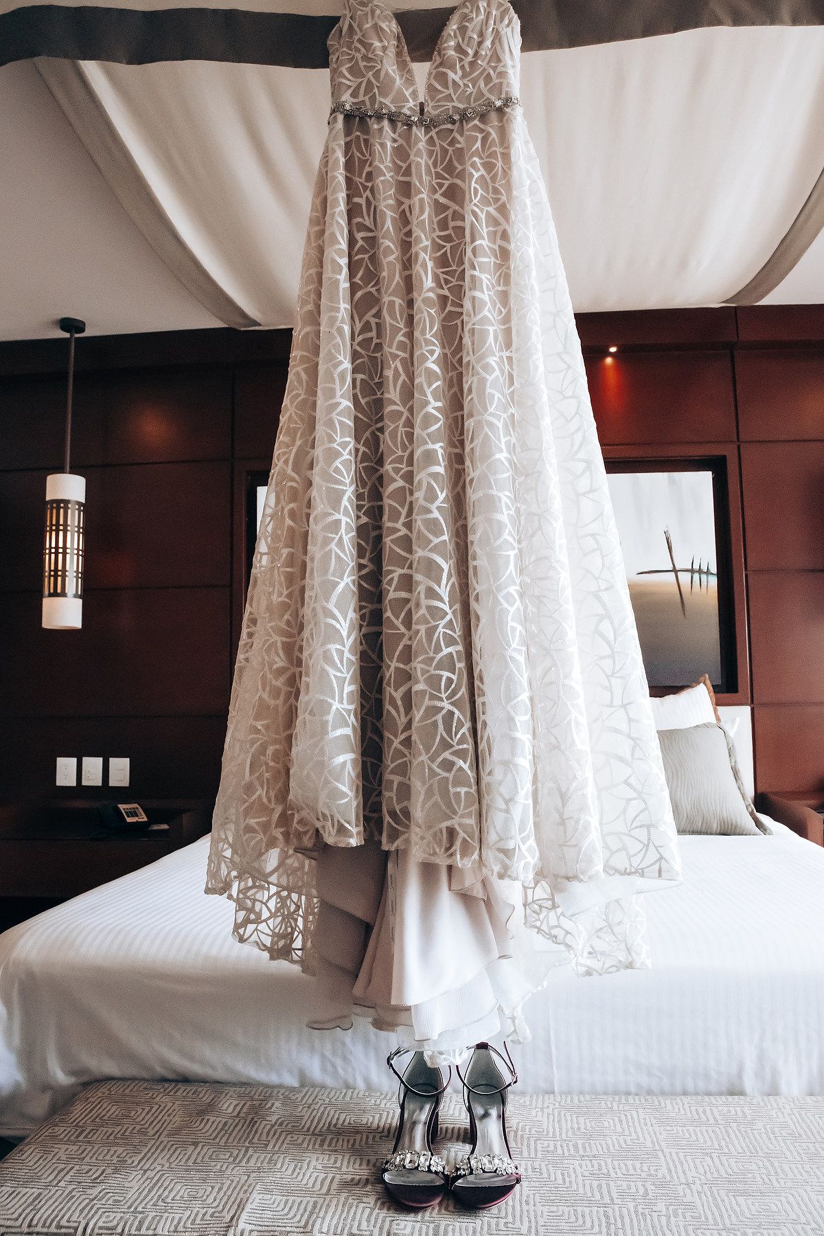 A white lace wedding dress hangs on a bed, with a pair of high heeled shoes visible on the floor.