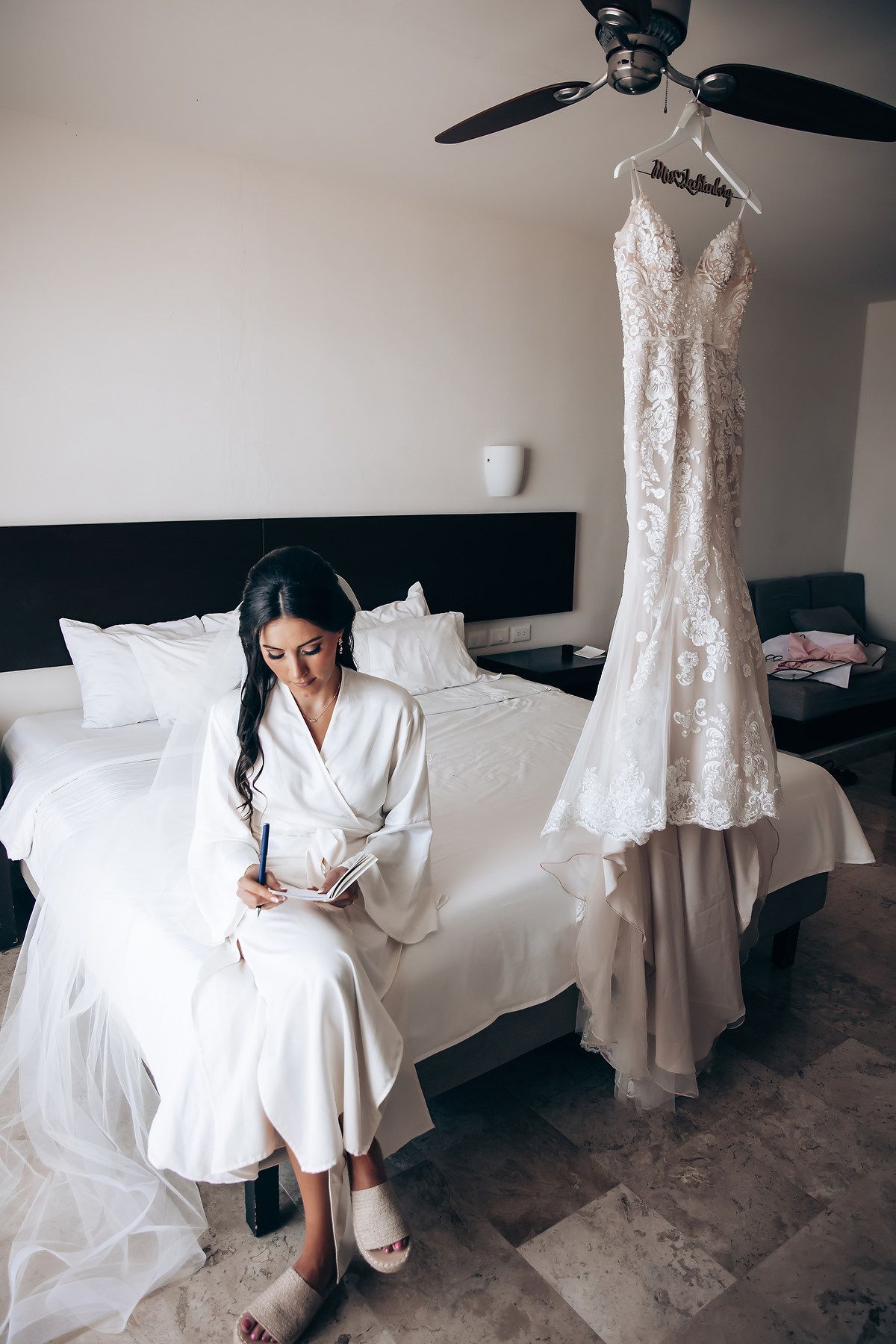 Wedding preparation scene with white dress hanging and person in robe writing at bedside.