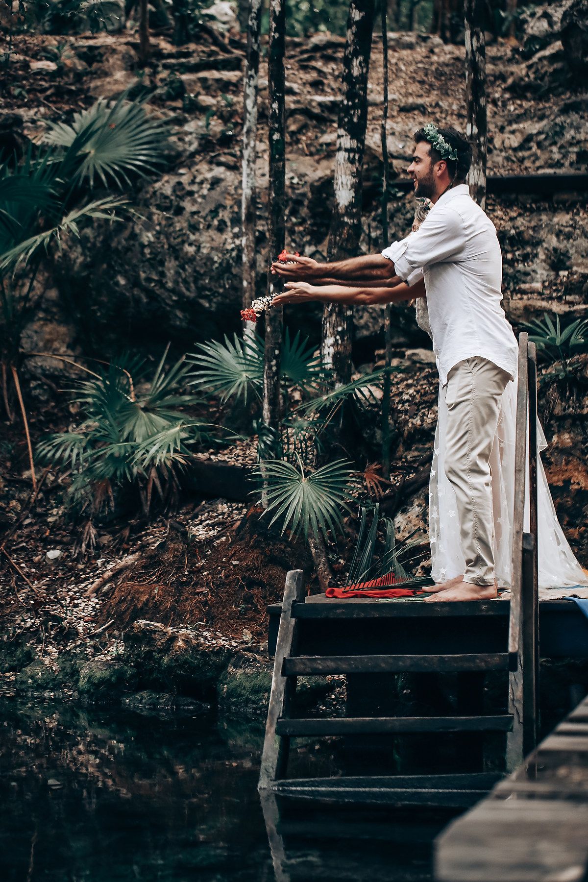 A person in white stands on wooden steps near a cenote wall with tropical palm fronds.