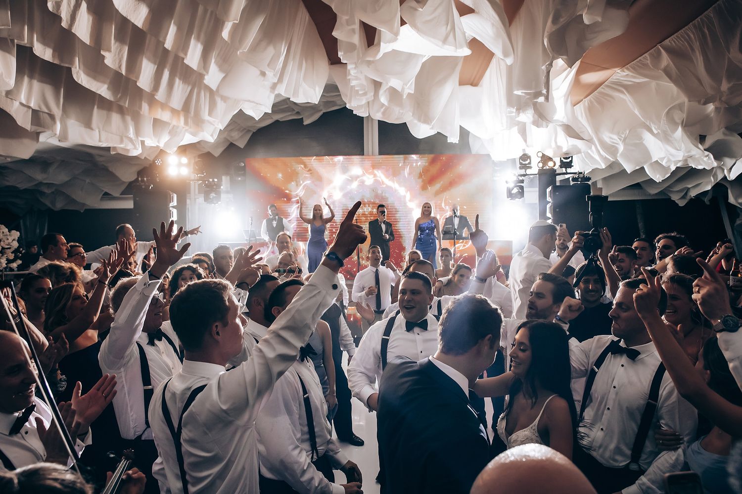 Large group of wedding guests dancing and celebrating under white ceiling draping.