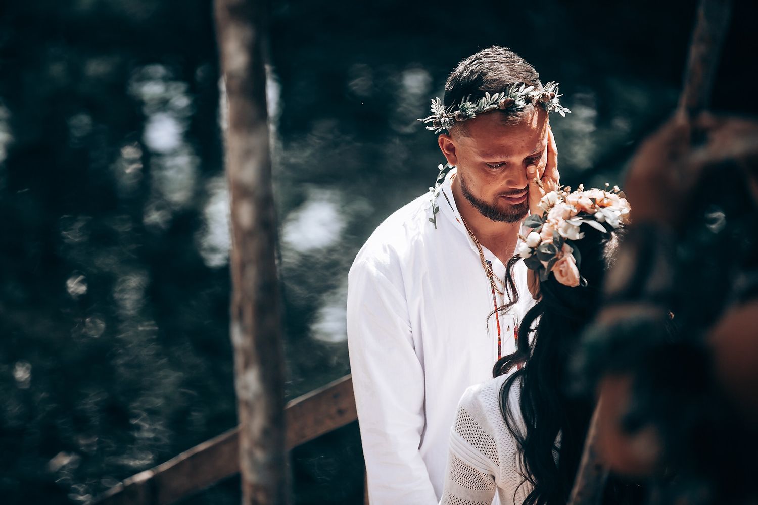 A person in white clothing wearing a floral crown stands near a tranquil forest pond.