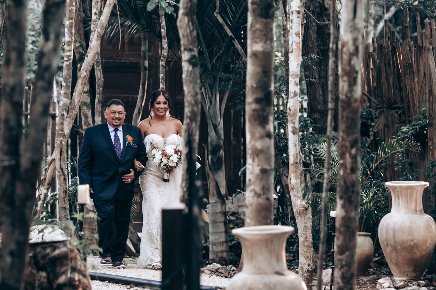 A wedding couple walks down a path lined with tall birch trees and decorative urns in a forest setting.