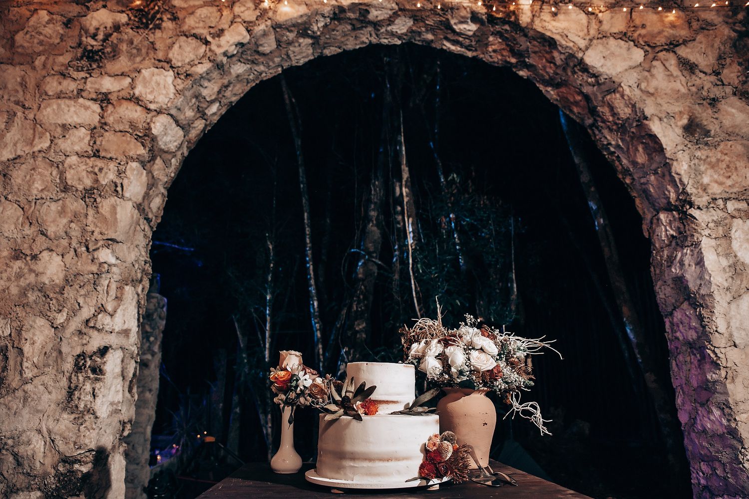 A rustic wedding cake display in a stone archway decorated with delicate floral arrangements.