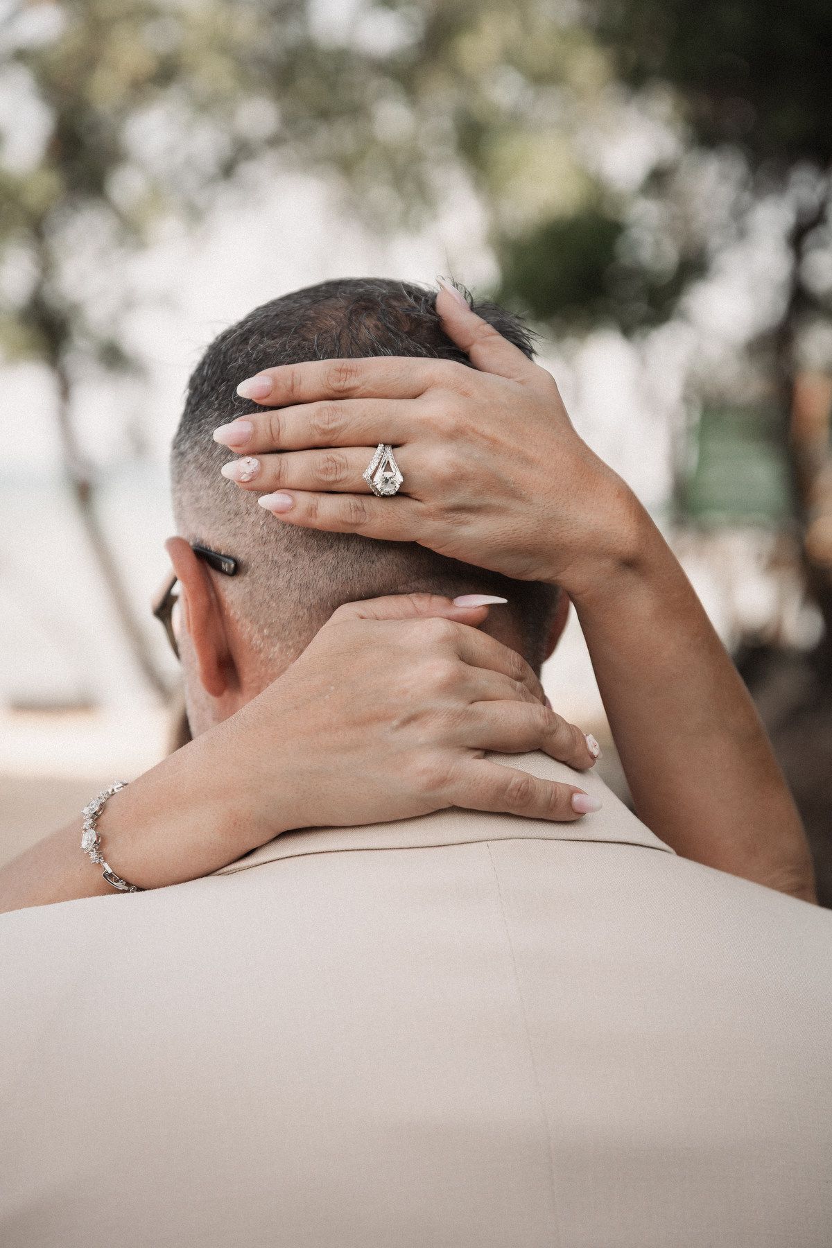 Bride's hands adorned with diamond ring and bracelet cradling groom's head at outdoor wedding.