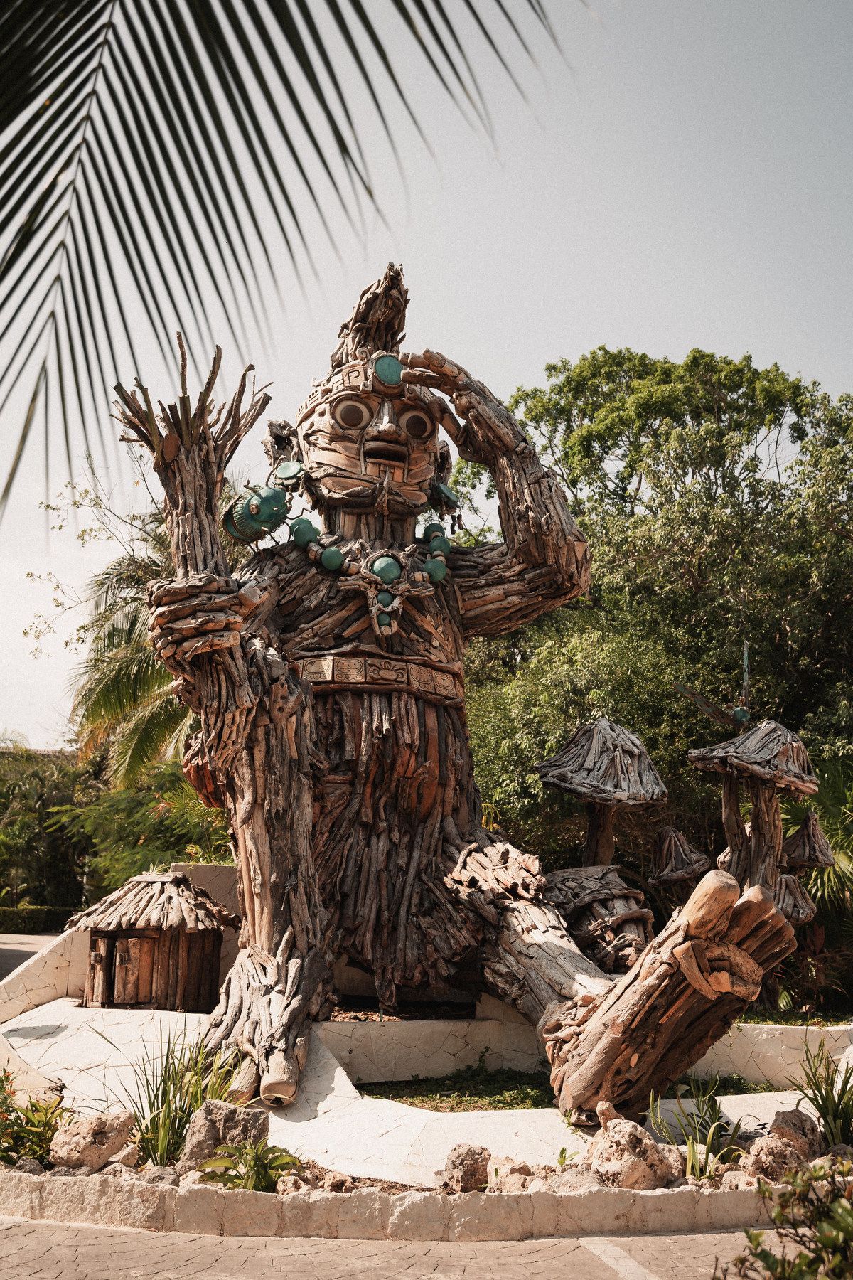 Large driftwood sculpture of a Mayan deity adorned with turquoise jewelry, surrounded by tropical trees and palm fronds.