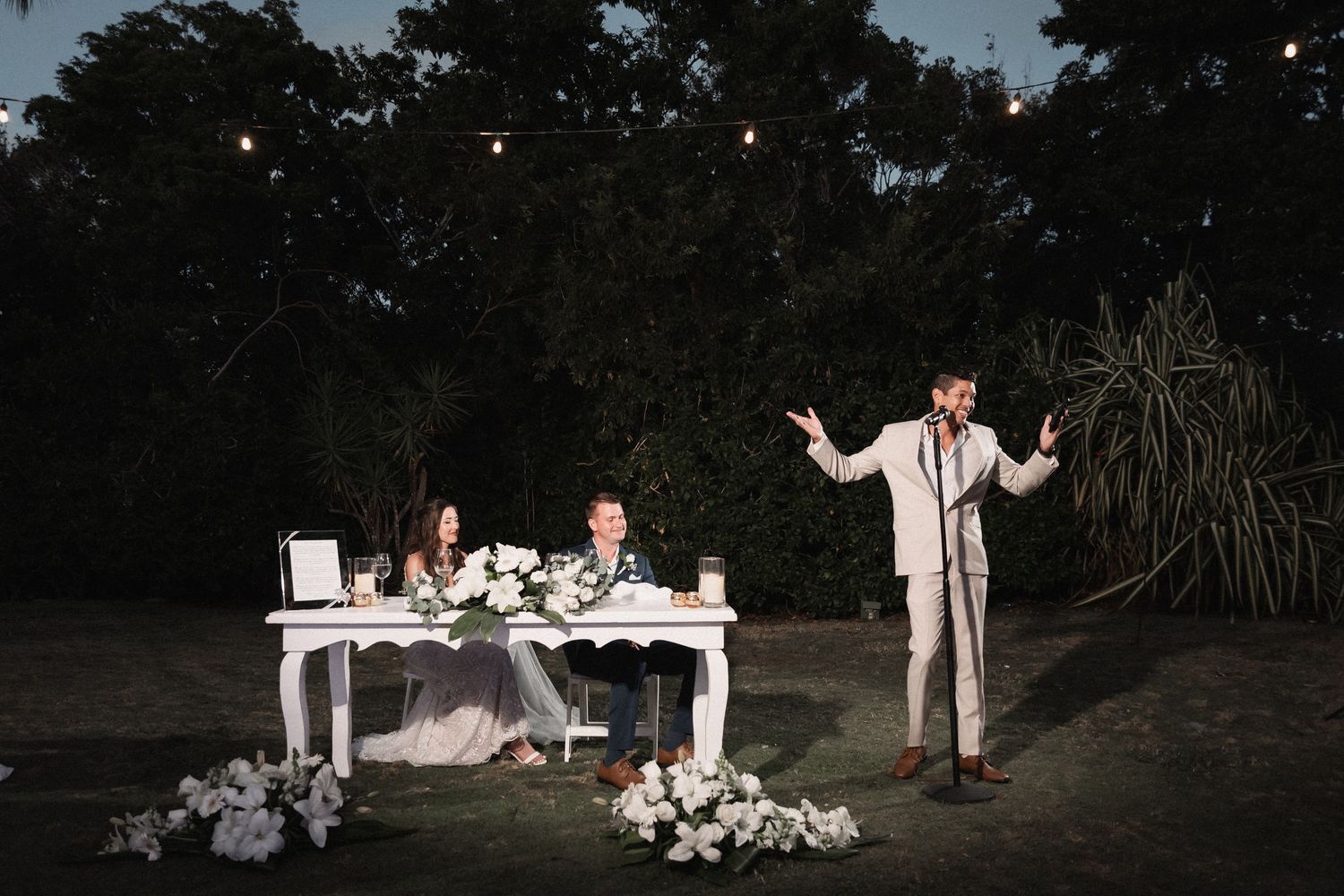 Best man gives speech at outdoor evening wedding reception with string lights overhead.