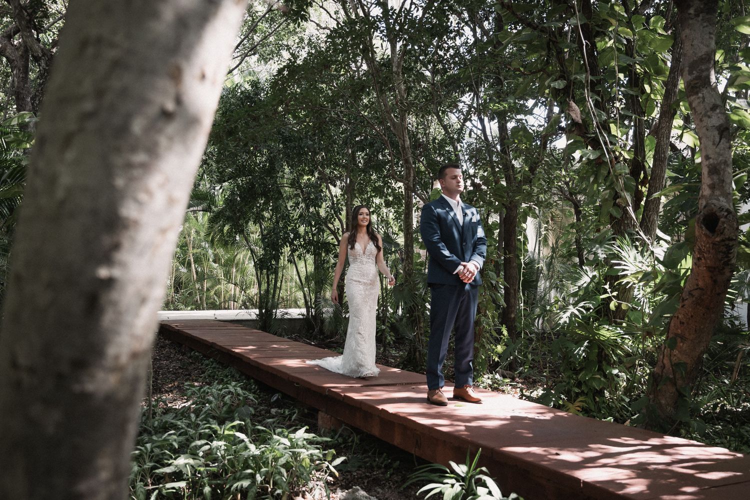 Bride and groom standing on wooden walkway surrounded by lush tropical greenery.