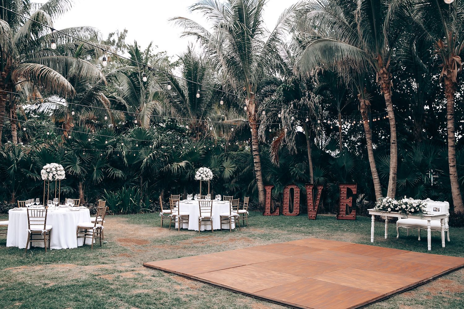 Outdoor wedding reception setup with dance floor and LOVE sign among tropical palm trees.