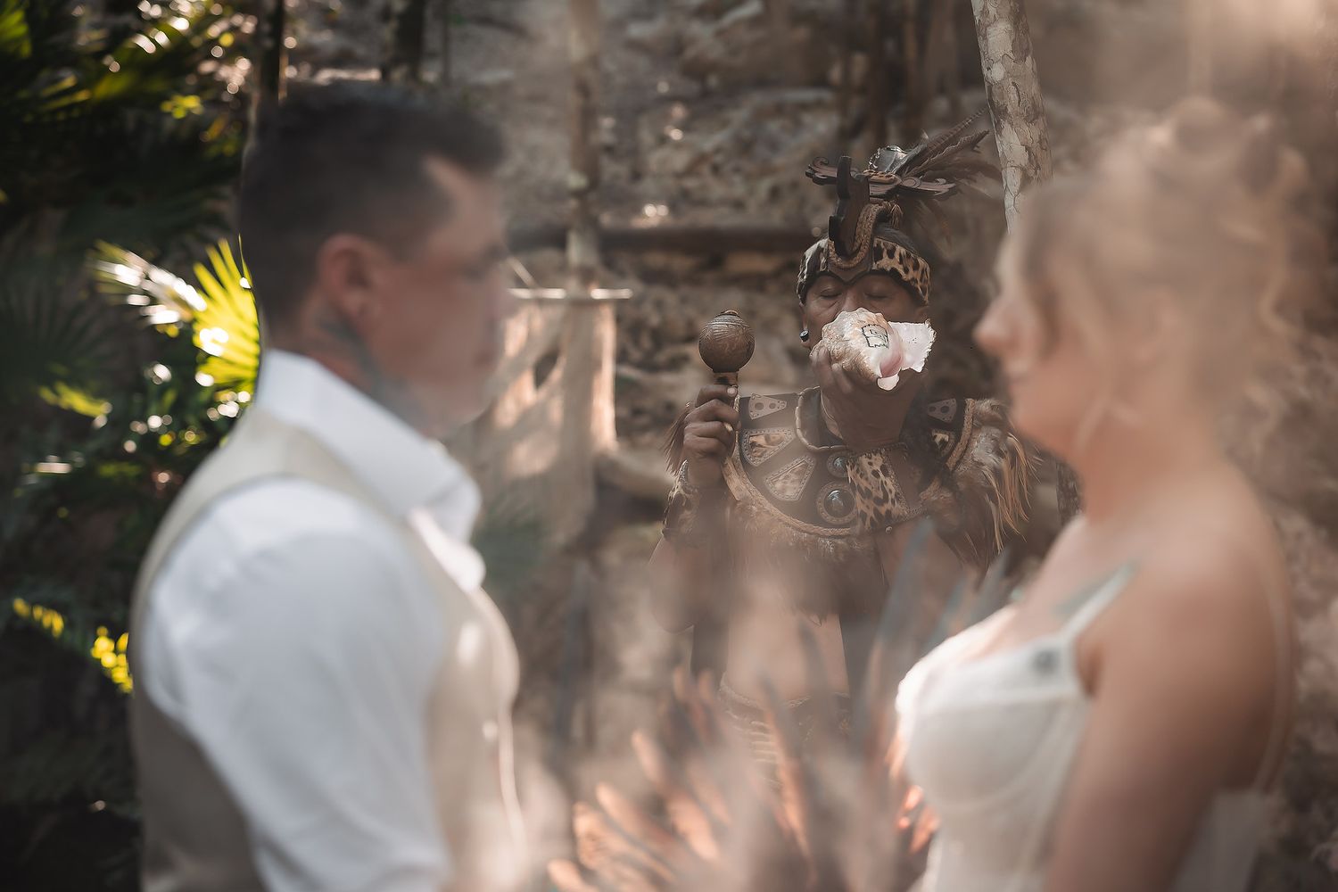 A couple stands before an ornate altar during an outdoor wedding ceremony surrounded by lush tropical greenery.
