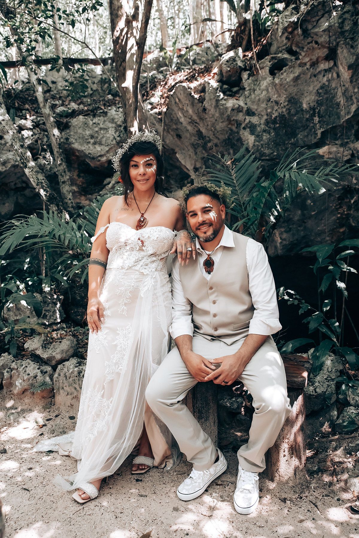 A couple sits together on stone steps in a garden setting wearing white and beige wedding attire.