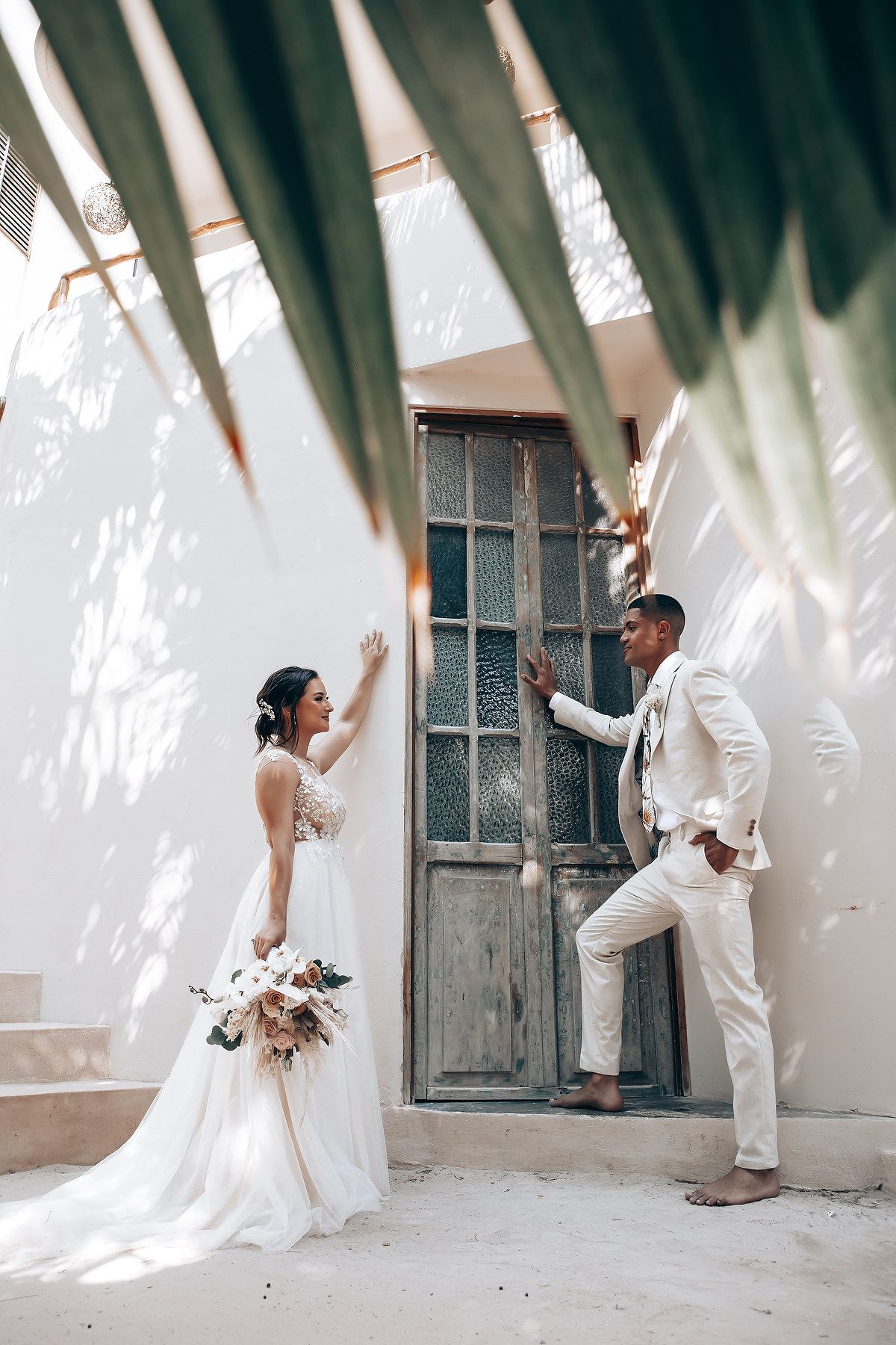 Bride and groom share playful moment through vintage doorway at tropical wedding venue.