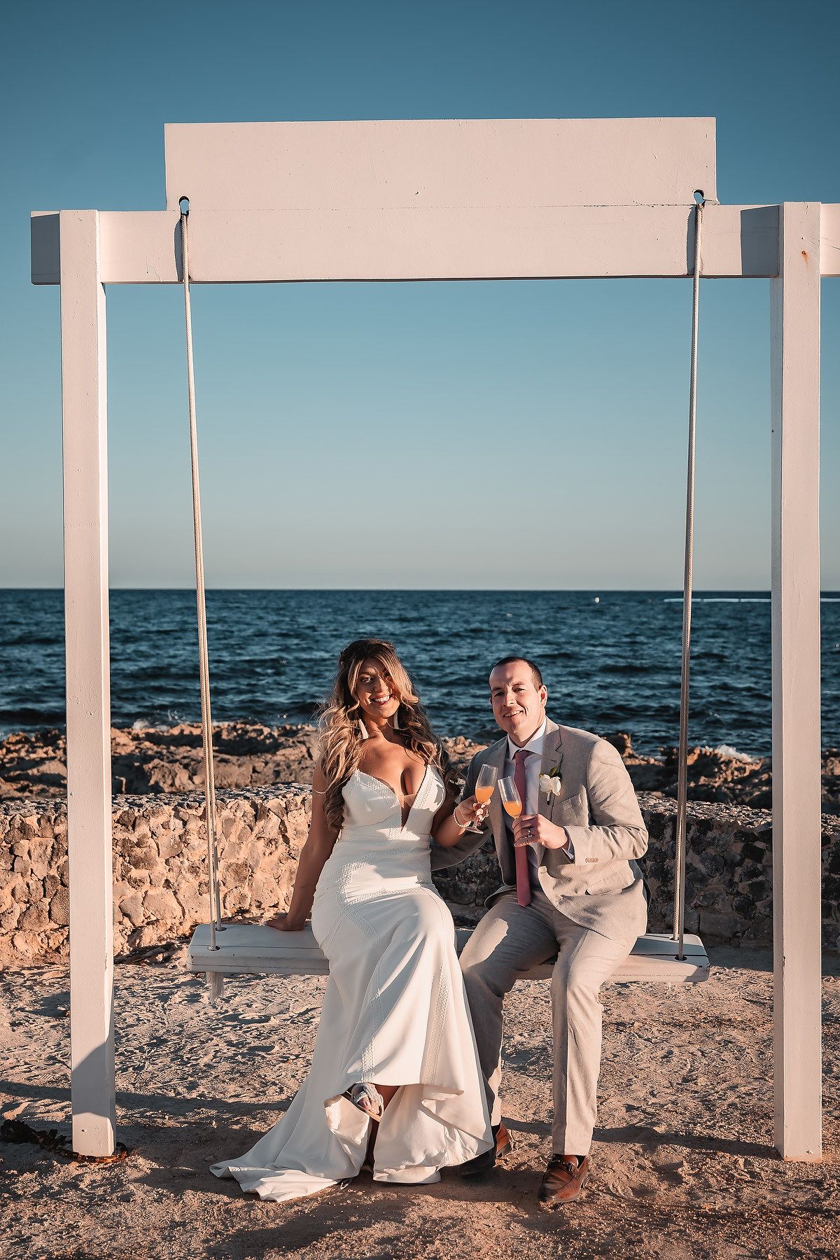 A couple in white attire poses playfully under white pillars by the ocean on a rocky beach.