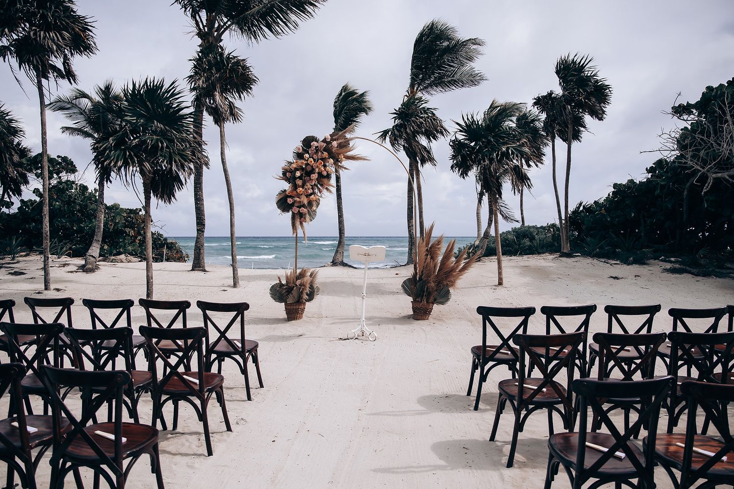 Wooden cross-back chairs line up on a tropical beach with palm trees and ocean views for an outdoor wedding ceremony.