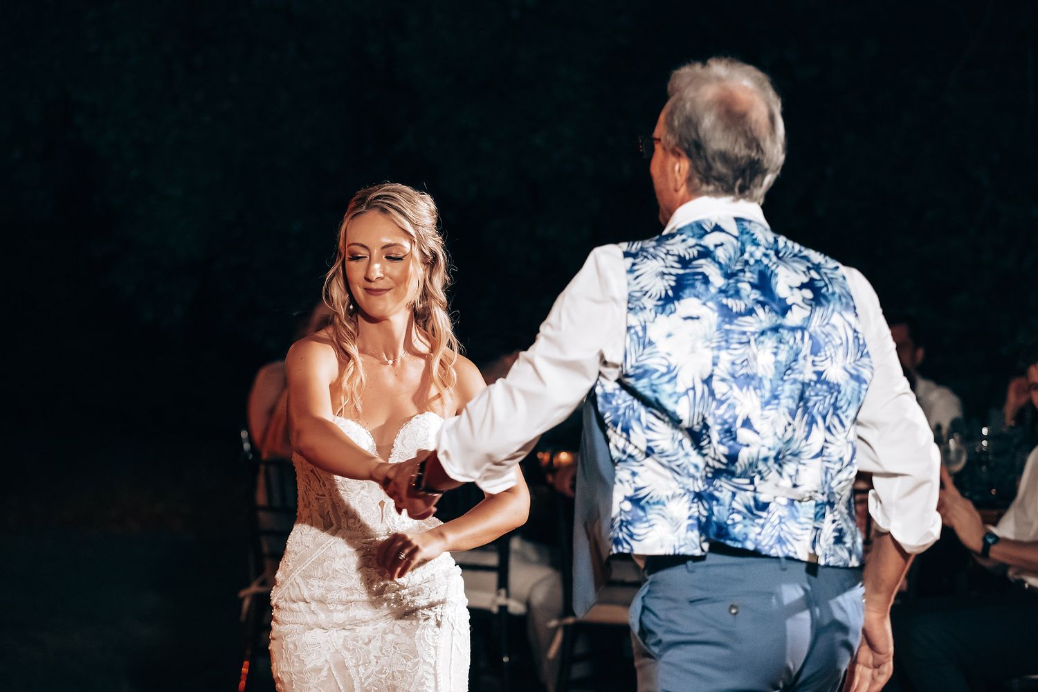 Wedding reception dance scene with bride in white dress and guest in blue floral shirt under dark lighting.