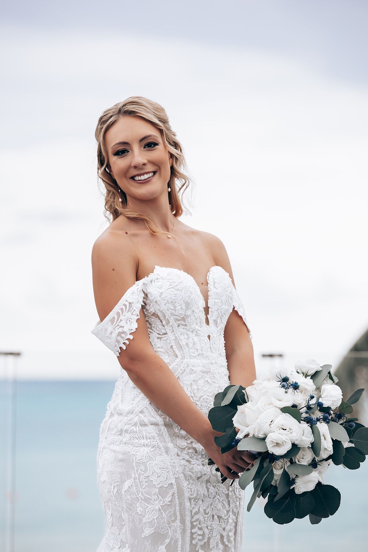 Bride in a white lace gown poses with a bouquet against an ocean backdrop.