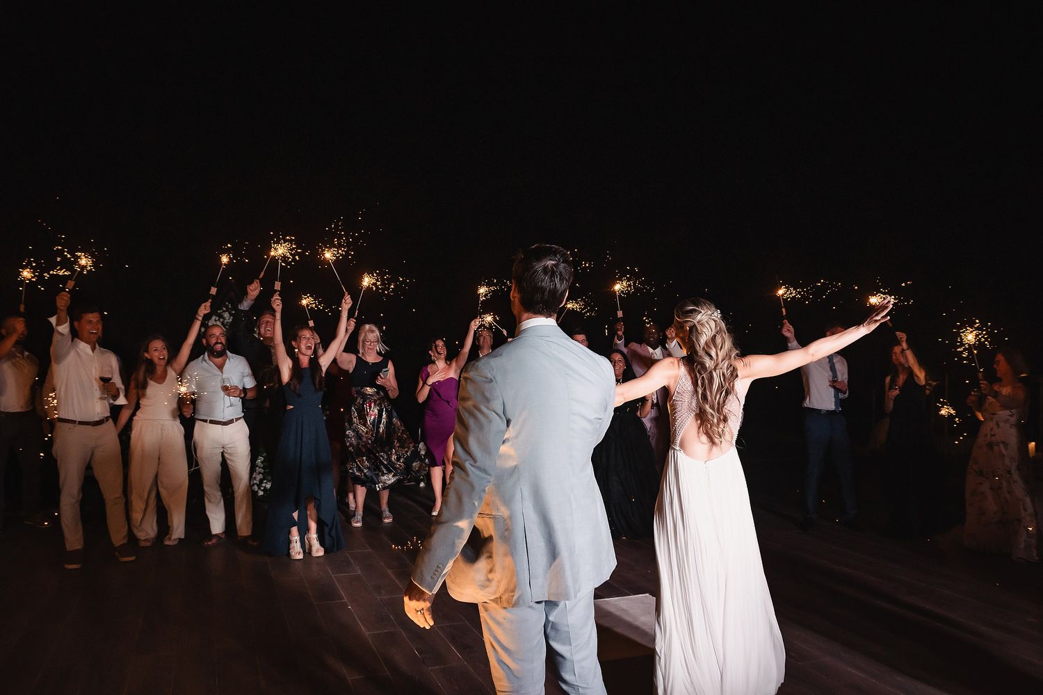 Wedding party celebrates on a dark outdoor deck while holding sparklers in the air.
