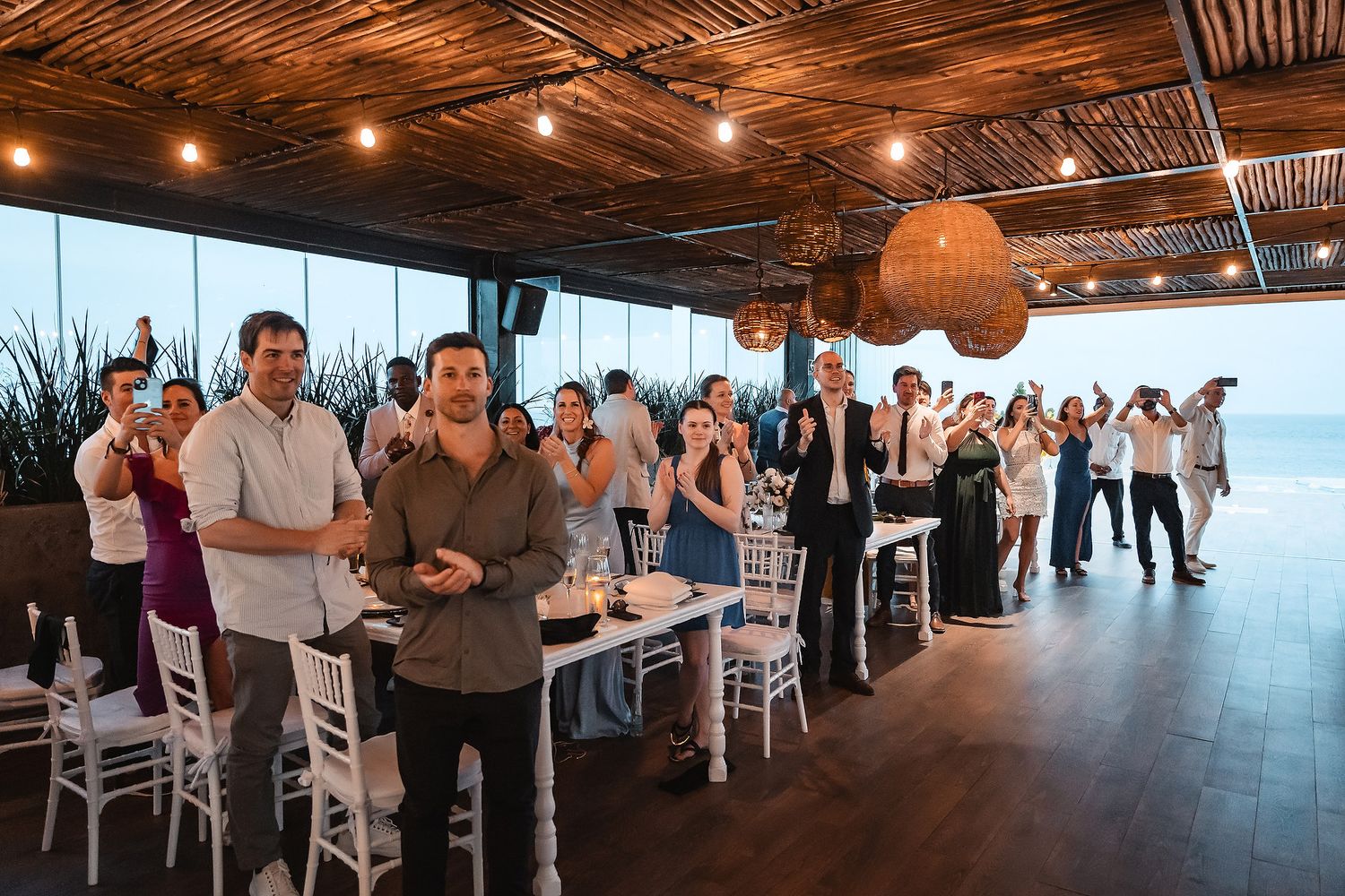 Wedding guests standing in modern venue with ocean views and string lights overhead.