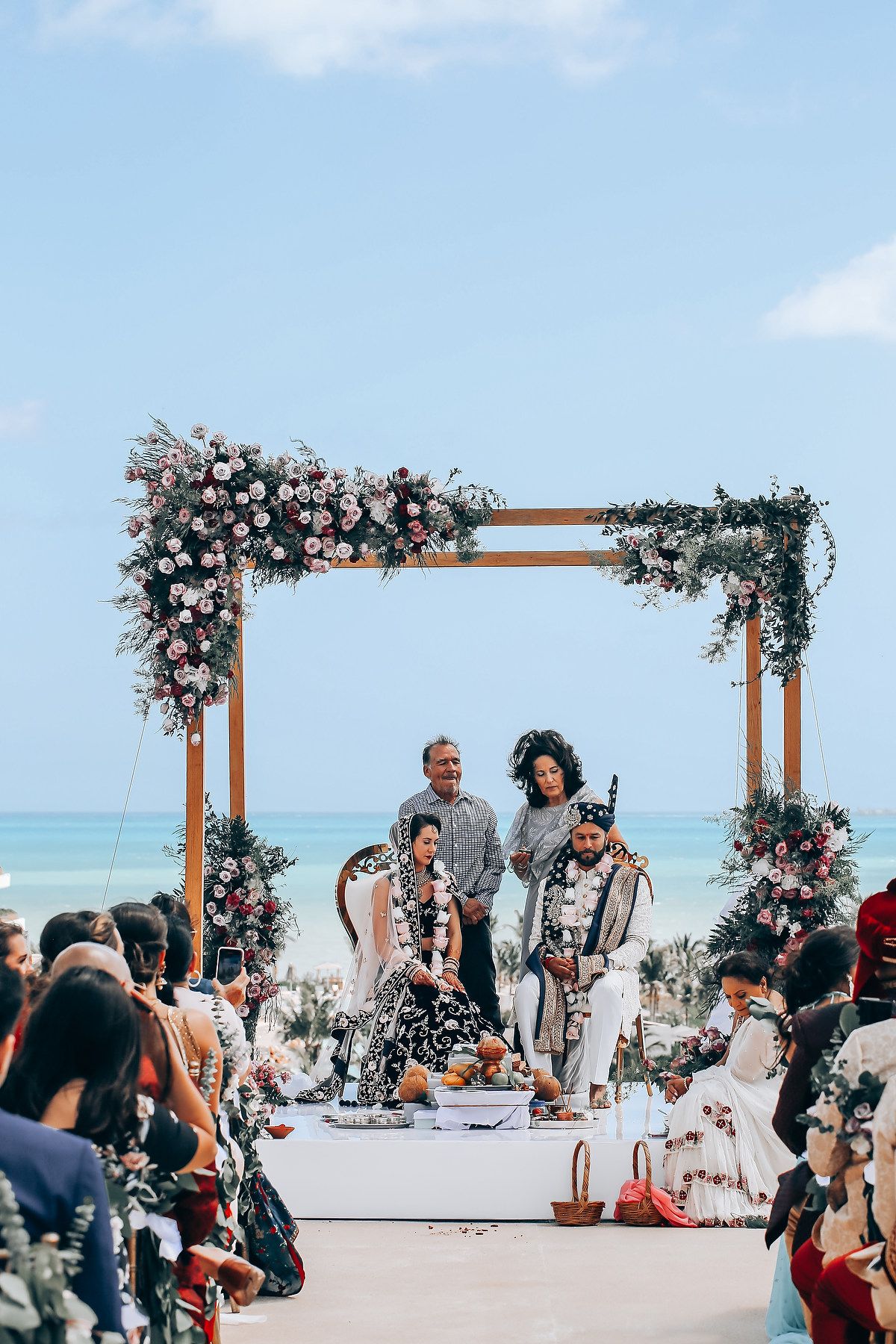 Wedding ceremony under wooden arch decorated with pink flowers against ocean backdrop.