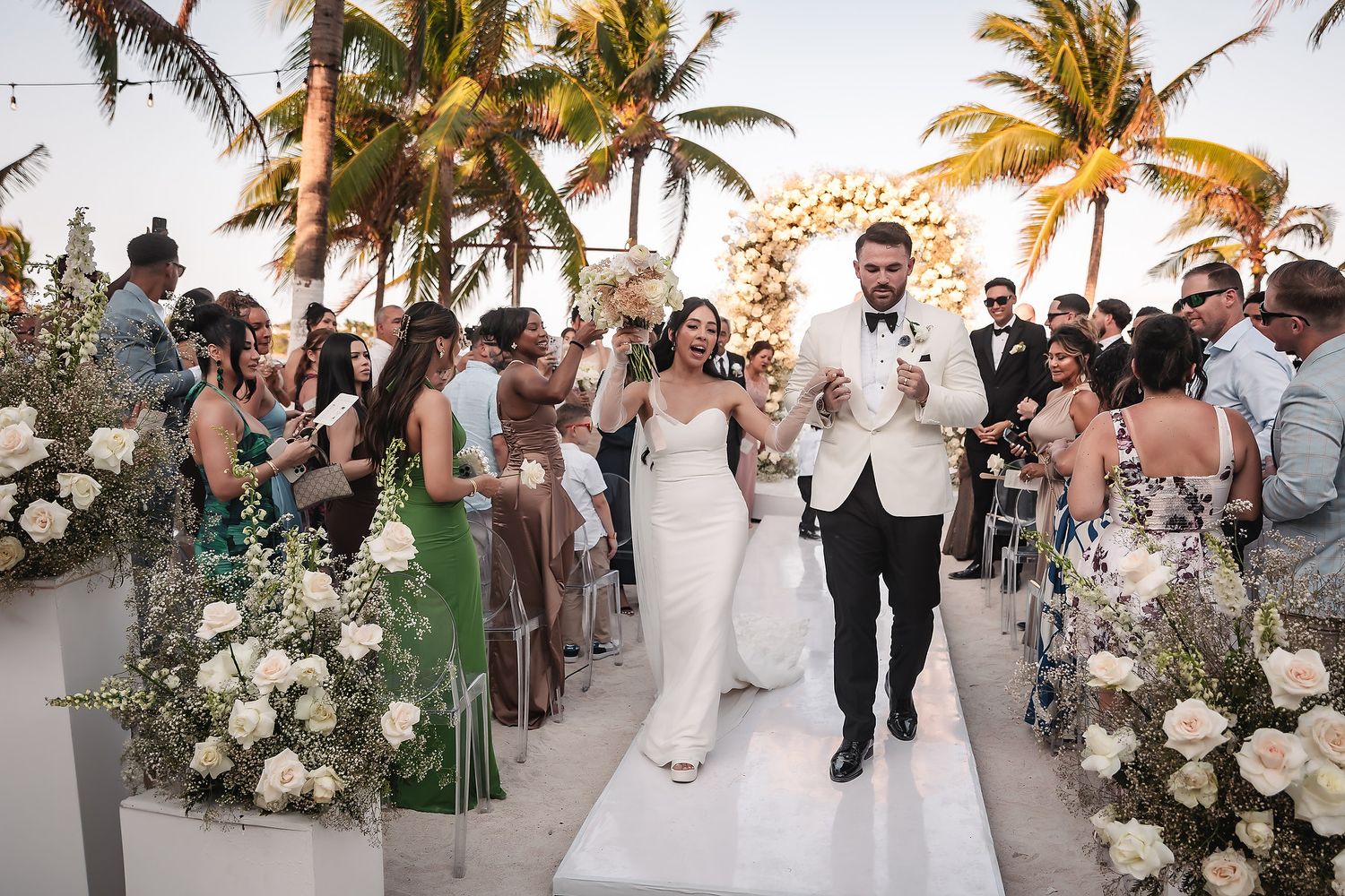 Couple walking down white aisle lined with floral arrangements and palm trees at beach wedding.