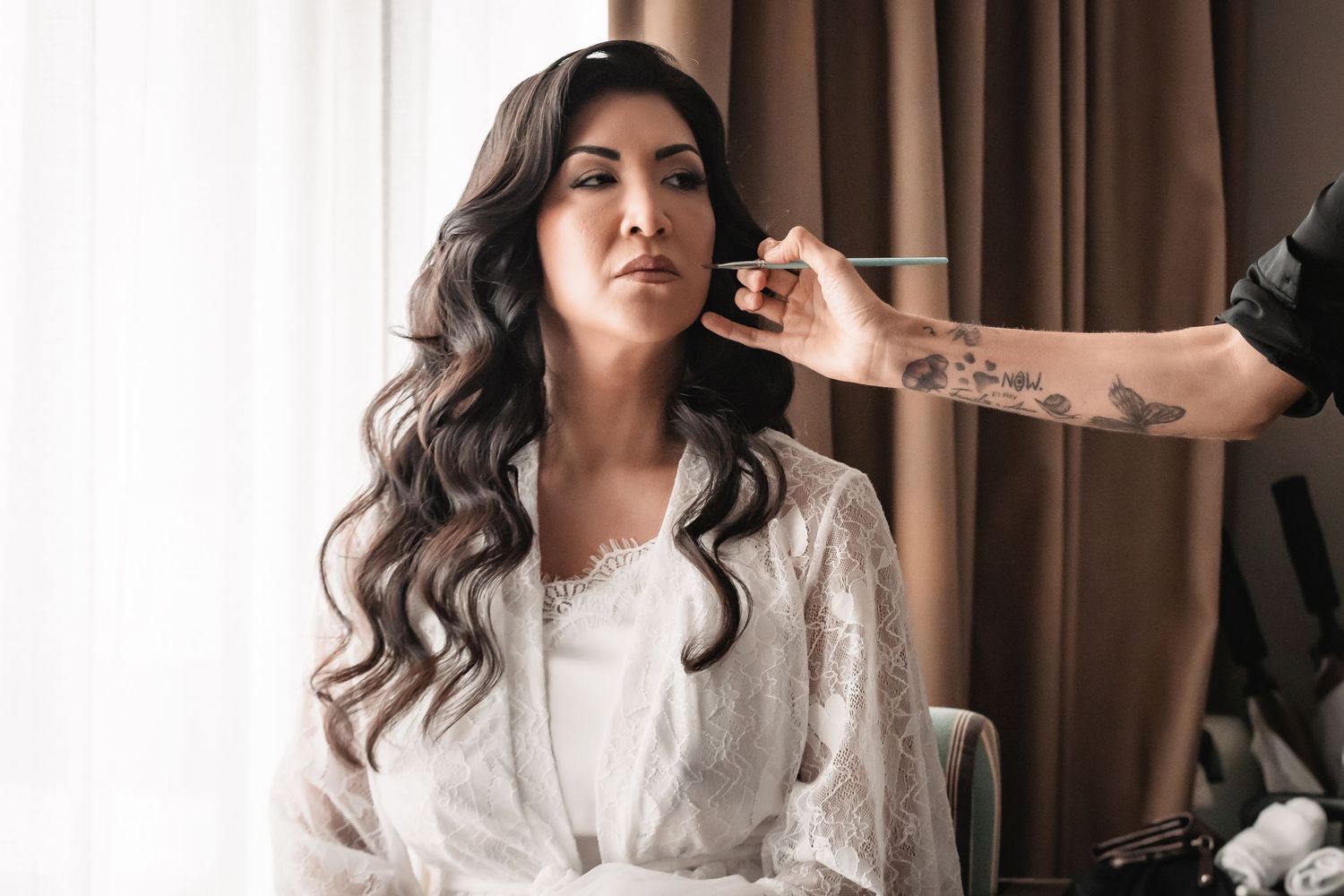 A makeup artist applies cosmetics to a person wearing a white lace robe during wedding preparations.