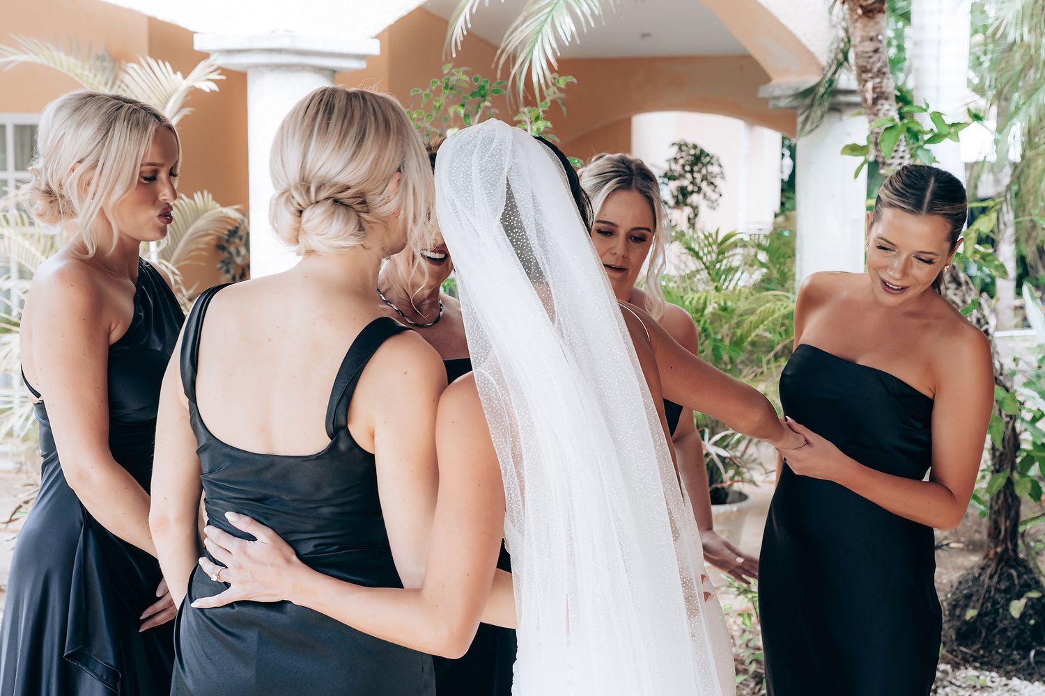 Bridesmaids in black dresses assist with wedding preparations in an outdoor covered walkway.