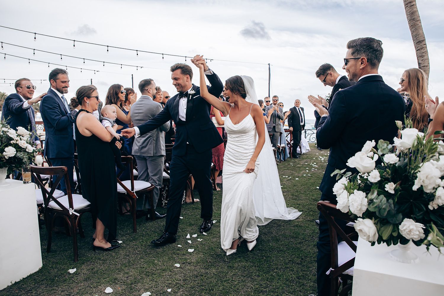 Newlyweds celebrate with guests during an outdoor seaside wedding ceremony with white floral decorations.