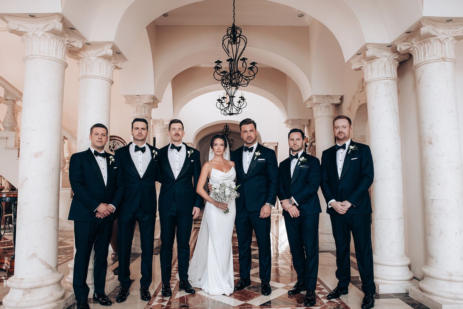 A wedding party in formal attire stands between elegant white columns in an ornate hallway.