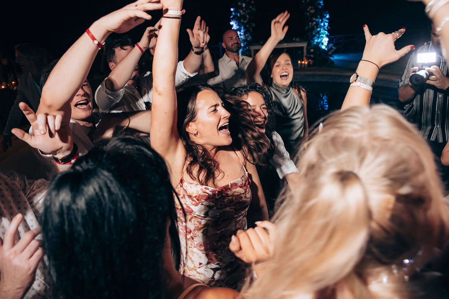 Crowd of people dancing with raised arms at a nightclub or concert venue with dark lighting and atmospheric effects.