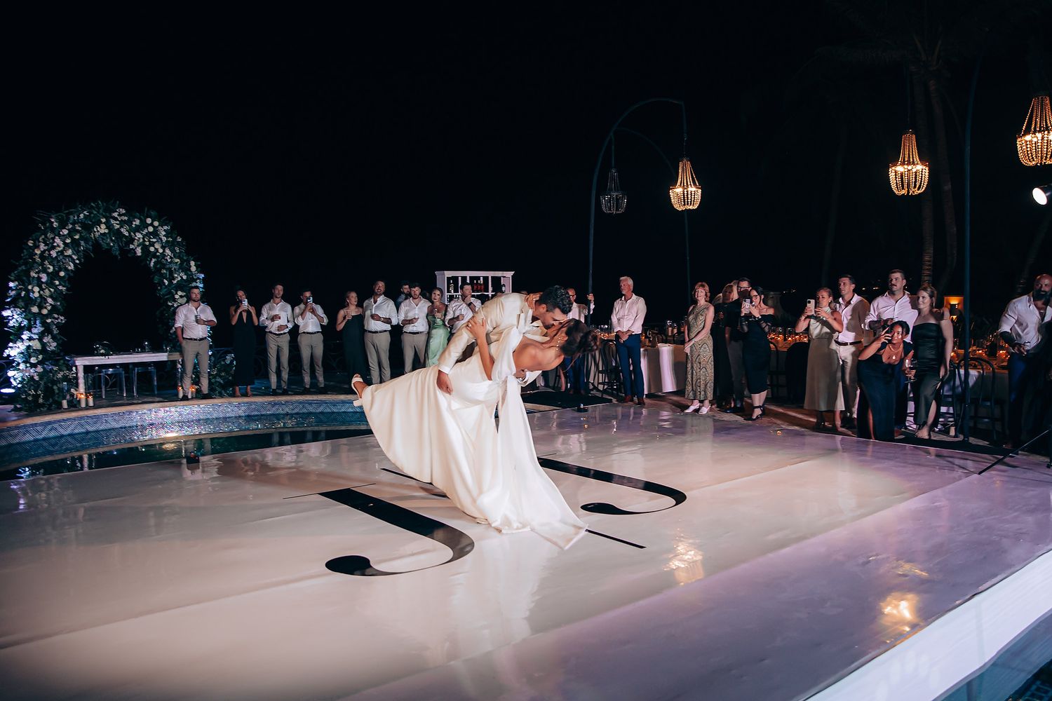 A couple performs their first dance on a white dance floor under string lights with guests watching at an outdoor reception.