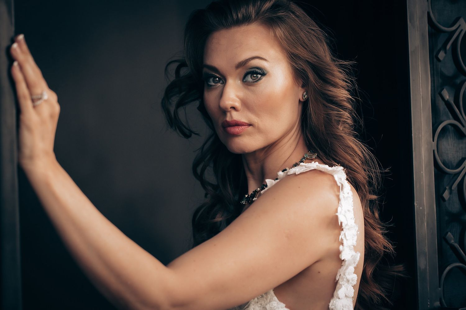 Woman with flowing dark hair in white sleeveless dress poses against dark wall in dramatic lighting.