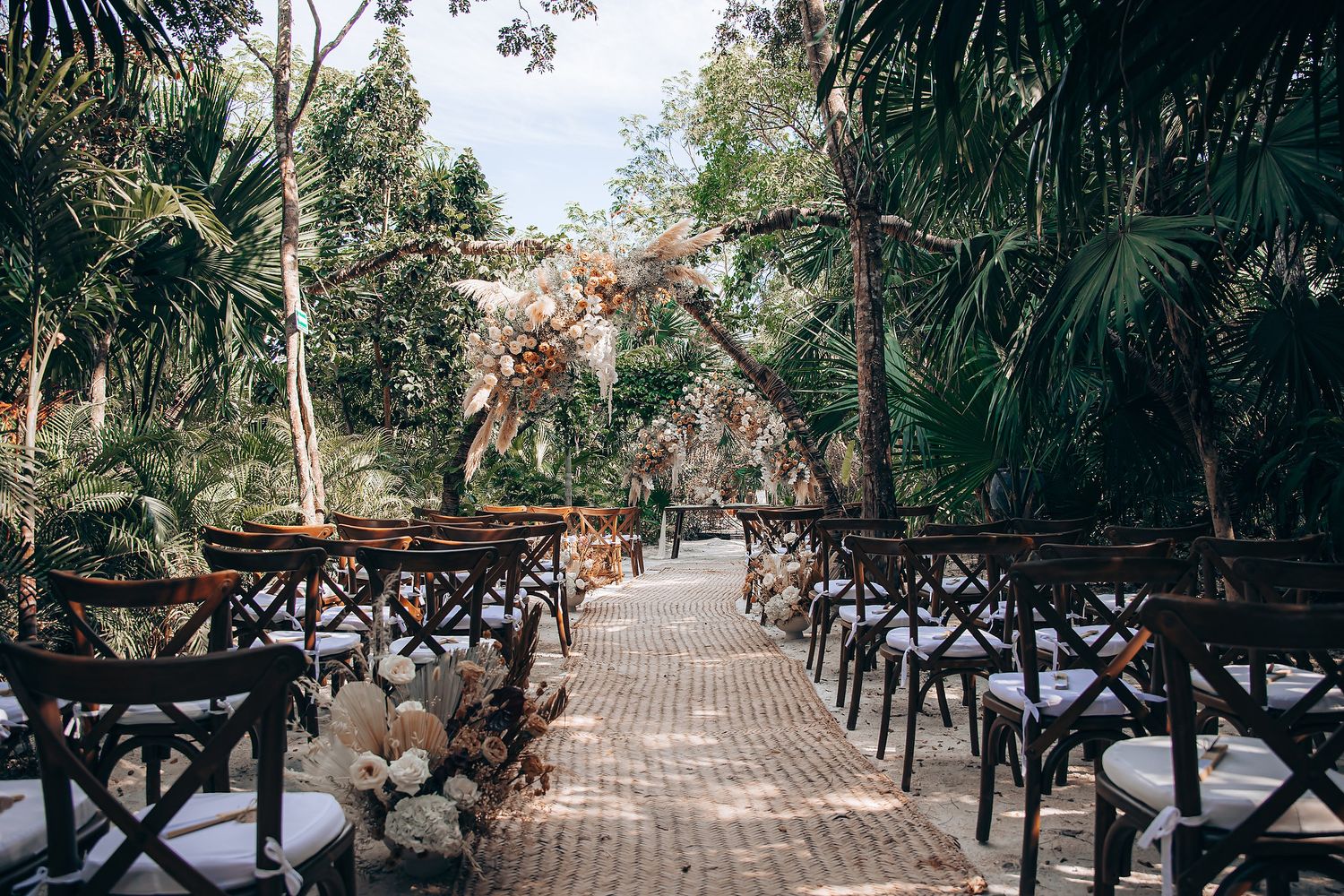 Rustic wooden chairs arranged in rows along a palm tree-lined pathway for an outdoor wedding ceremony.