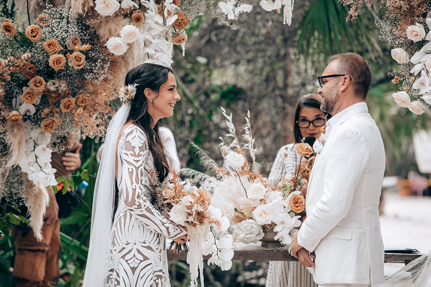 Couple in elegant white wedding attire poses together surrounded by delicate floral decorations in an outdoor setting.