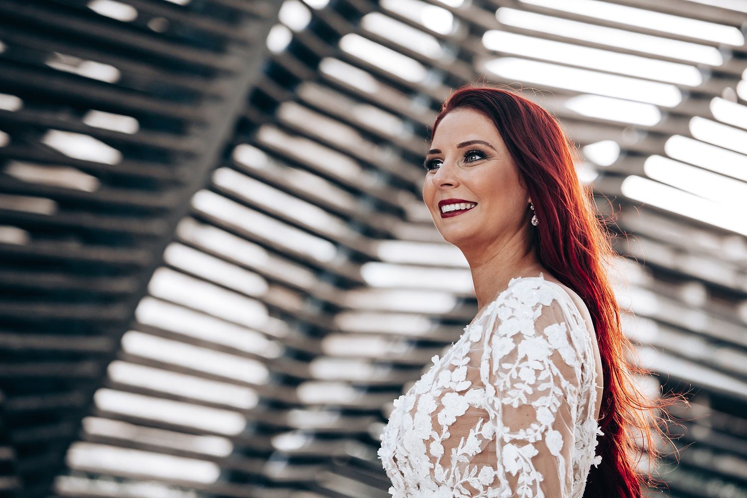 Bride in lace dress poses under modern architectural structure with natural lighting.
