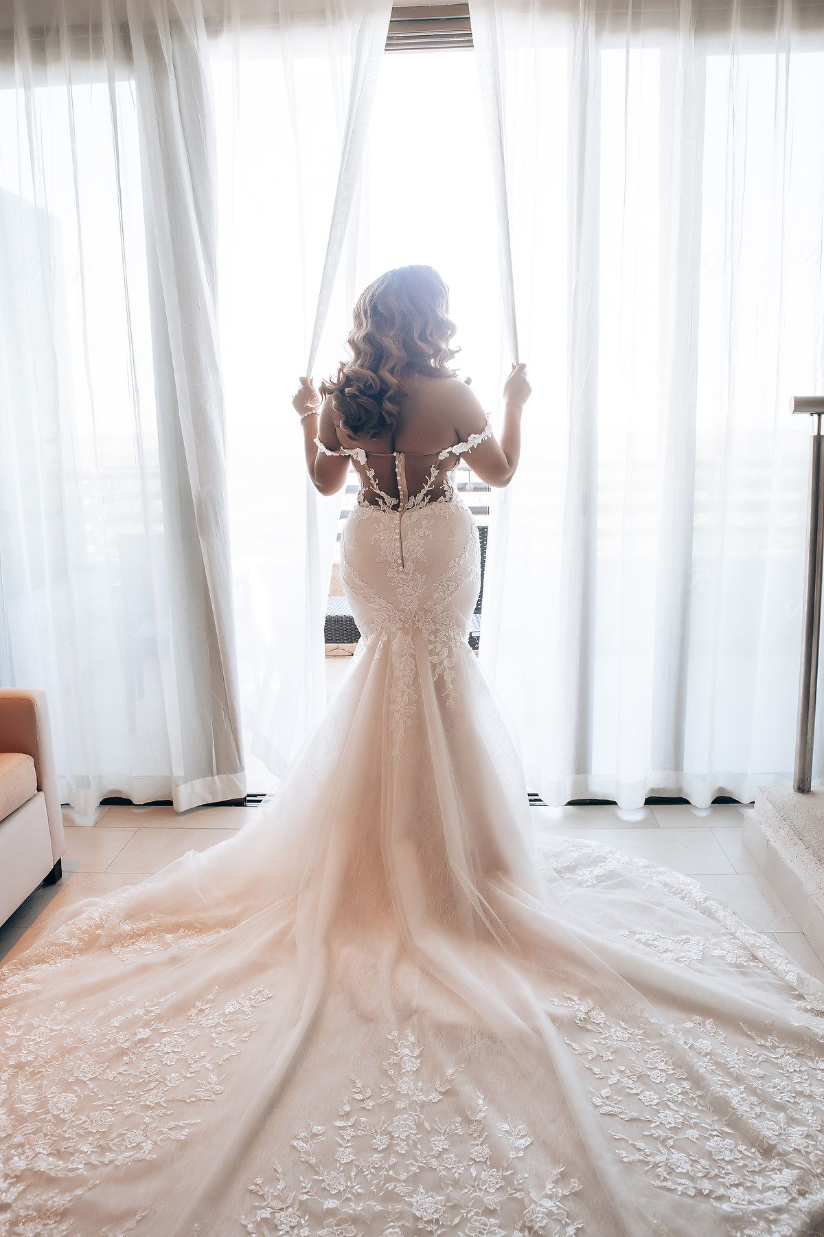 Back view of wedding dress with long train and delicate lace details against white curtains.