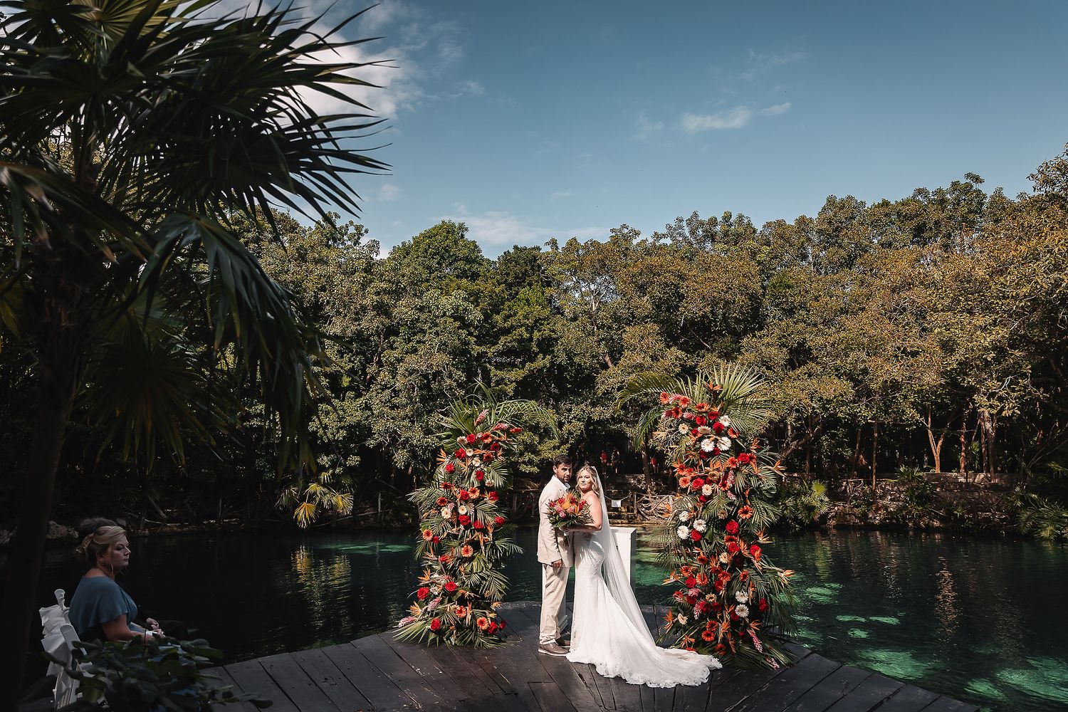 Waterfront wedding ceremony with red floral arches and palm trees under blue skies.