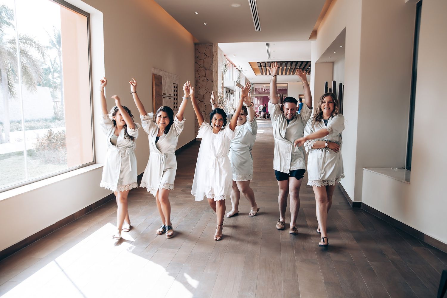 Wedding party members in white dresses and casual attire walk down a modern hallway celebrating.