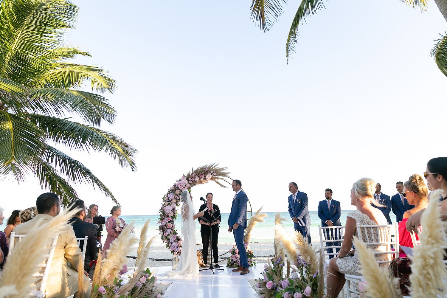 Wedding ceremony guests gather under swaying palm trees on a tropical beach with white chairs and ocean views.