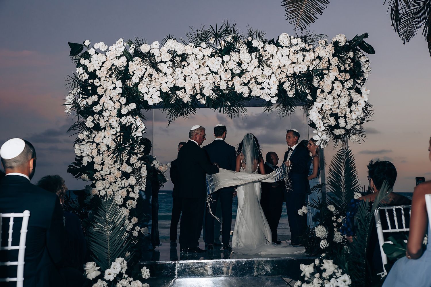Sunset wedding ceremony beneath an ornate white floral arch with ocean views.