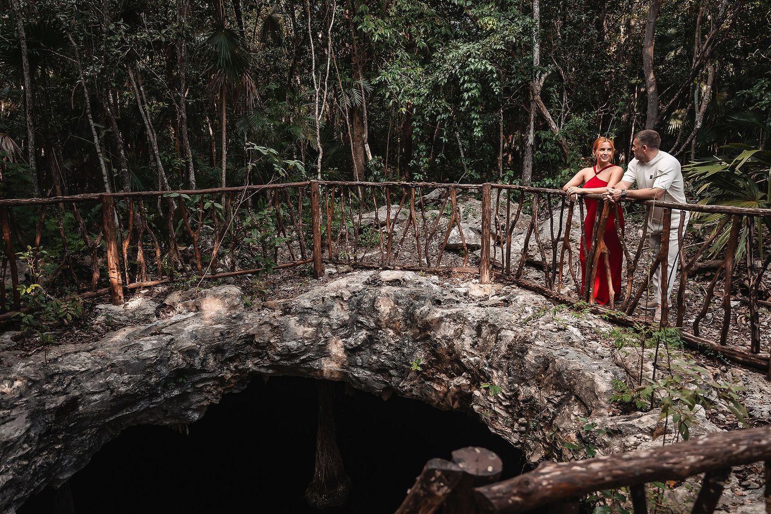 A couple stands on a stone bridge overlooking a cenote in a tropical forest.
