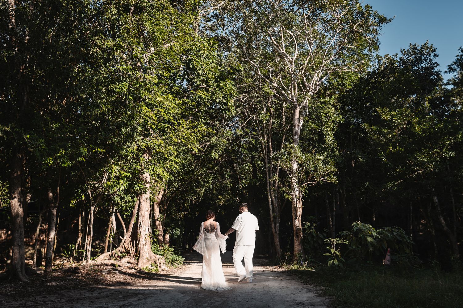 A couple in wedding attire walks along a sunlit forest path surrounded by lush green trees.