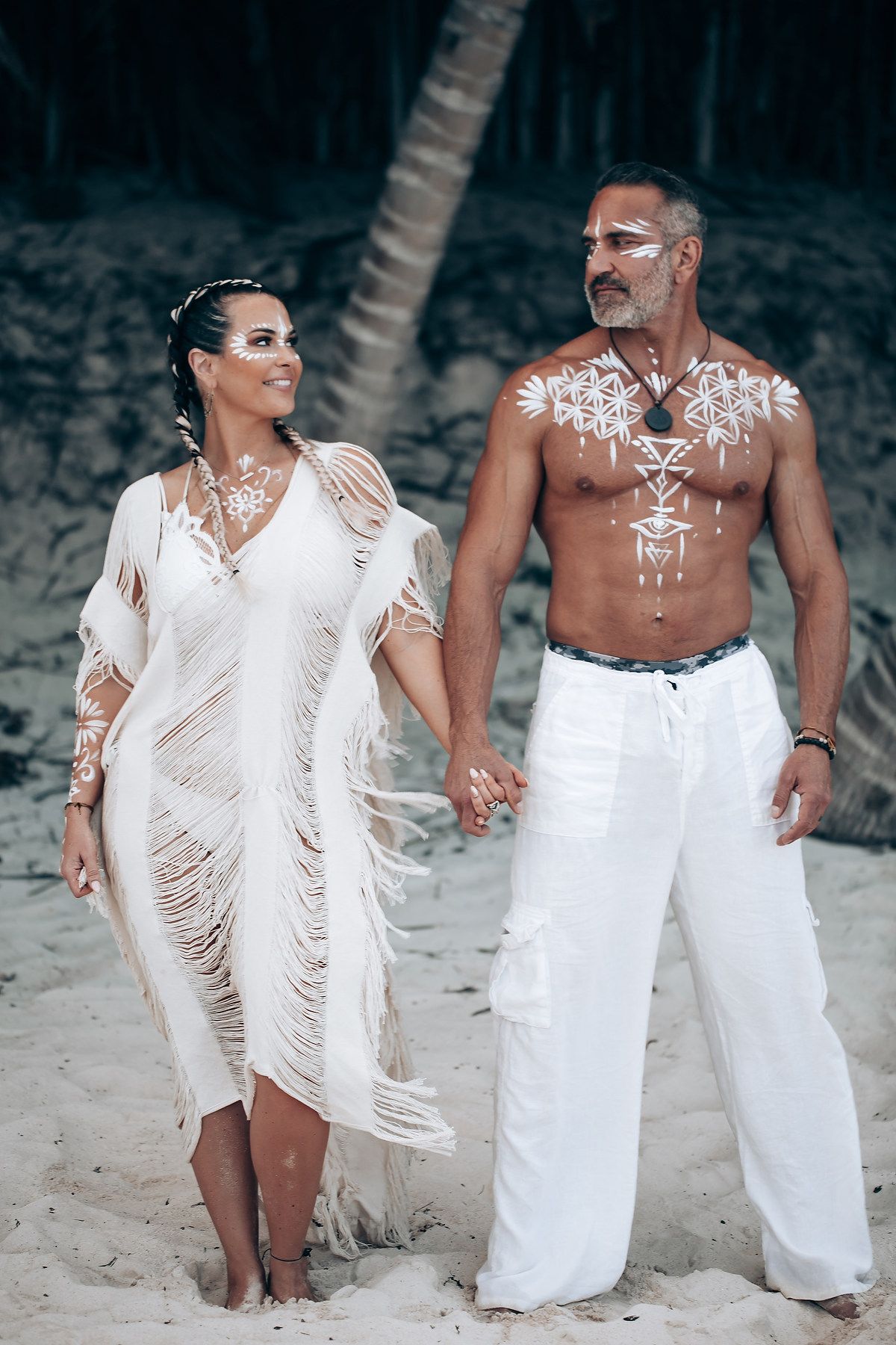 A couple in white beachwear with decorative body paint poses against a rustic backdrop.