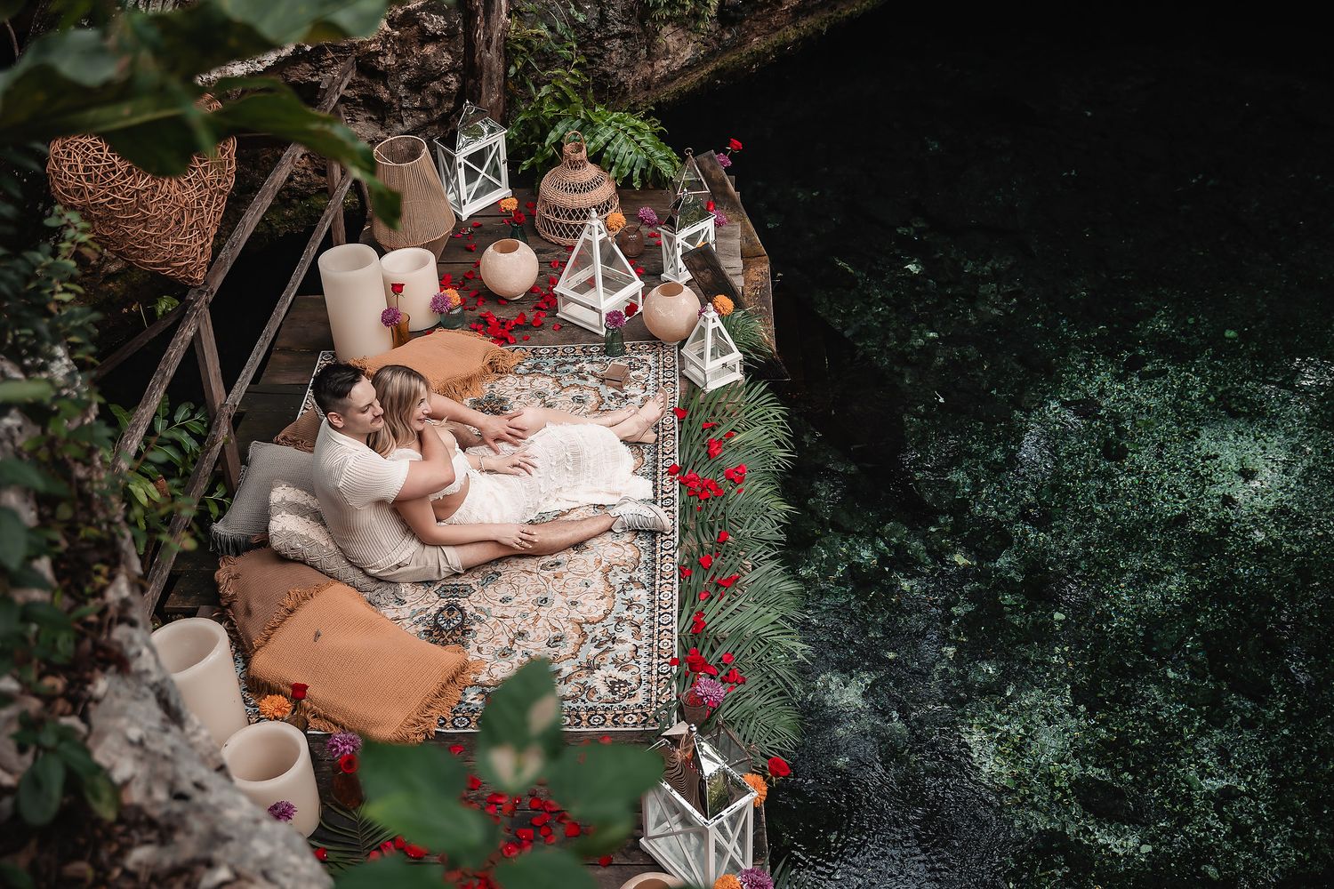 Romantic outdoor setup with blankets, pillows and candles next to a dark reflective pool.