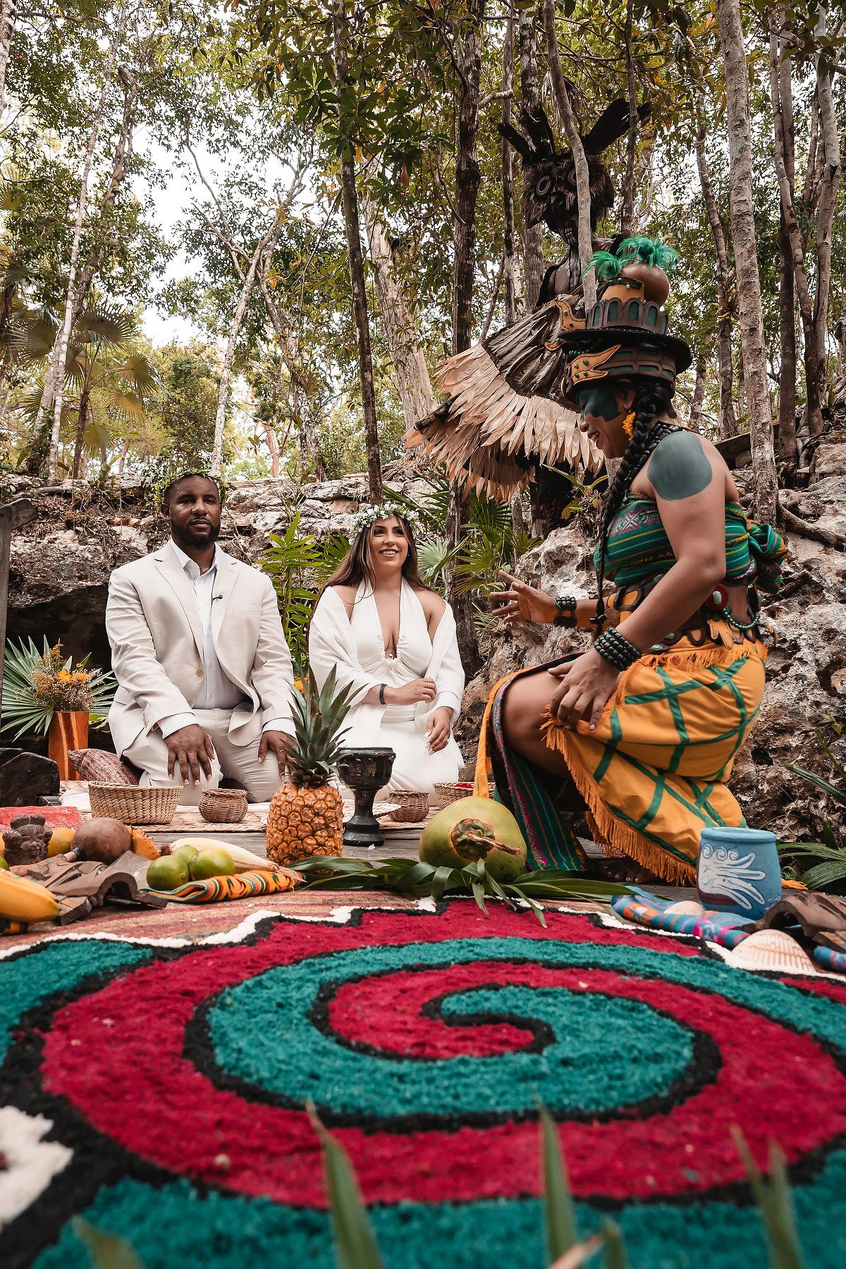 Traditional ceremony in a forest setting with colorful rugs and tropical fruits displayed on the ground.