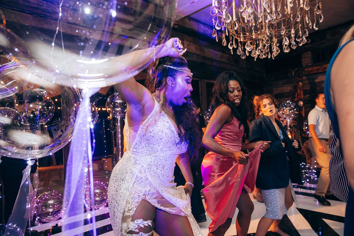 People dancing and celebrating at a wedding reception under purple lighting and crystal chandeliers.