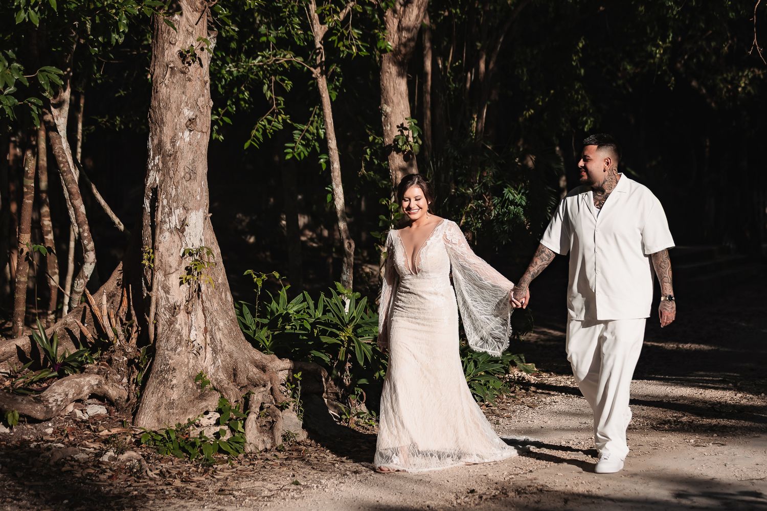 A couple strolling together on a tree-lined path in white wedding attire.