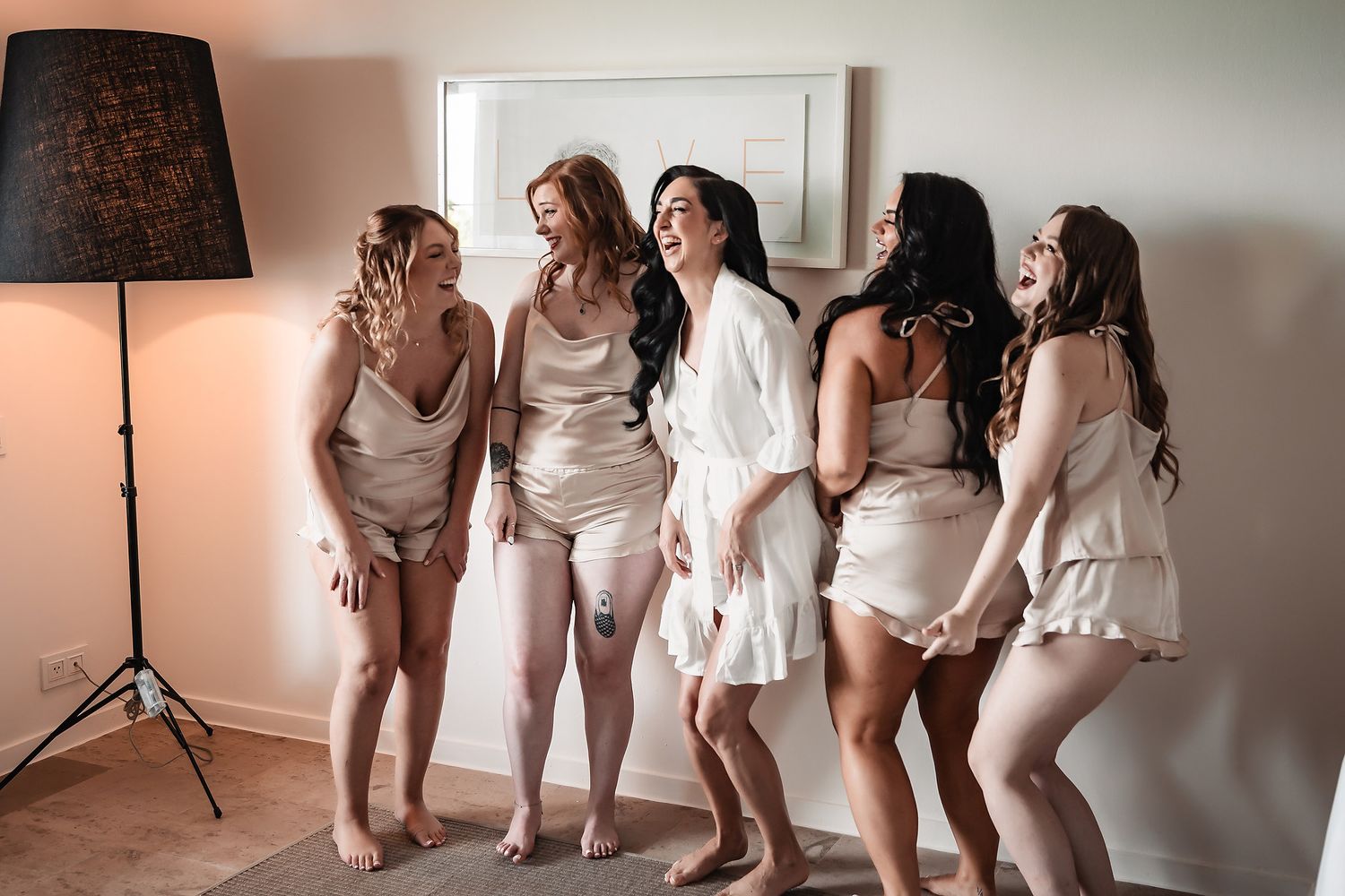 A group poses together in white dresses and robes while sitting on a white couch during a wedding preparation.